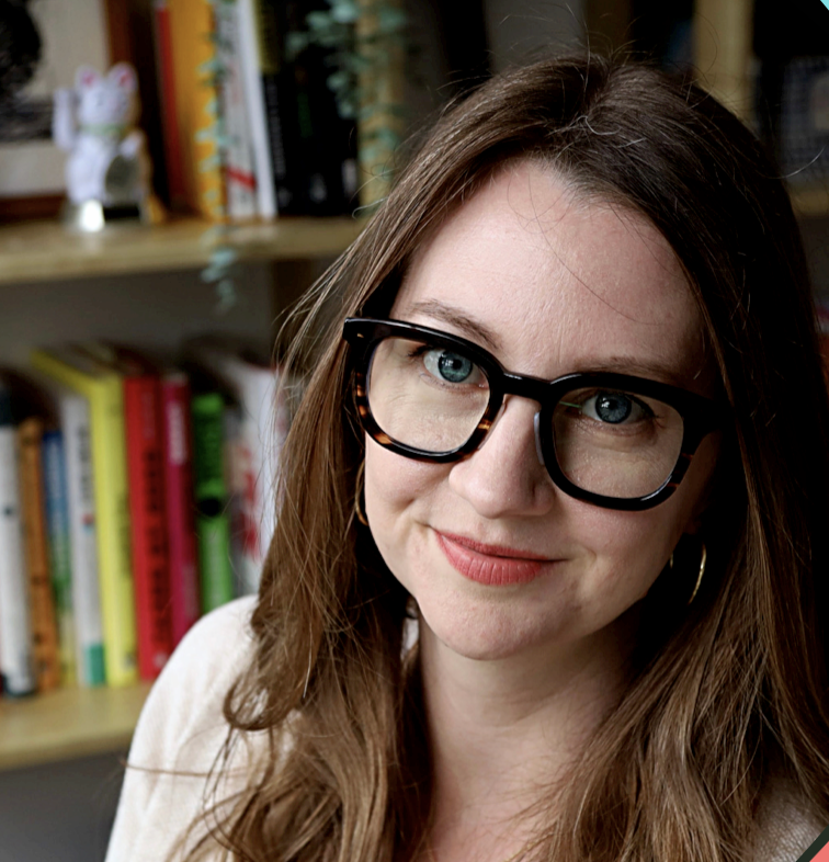 Dana Publicover smiles at the camera. She is a white woman with light brown hair and glasses with hesavy black frames. She is sitting in front of a bookcase.