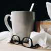 A white mug with a spoon in I, surrounded by a pair of glasses and tissues
